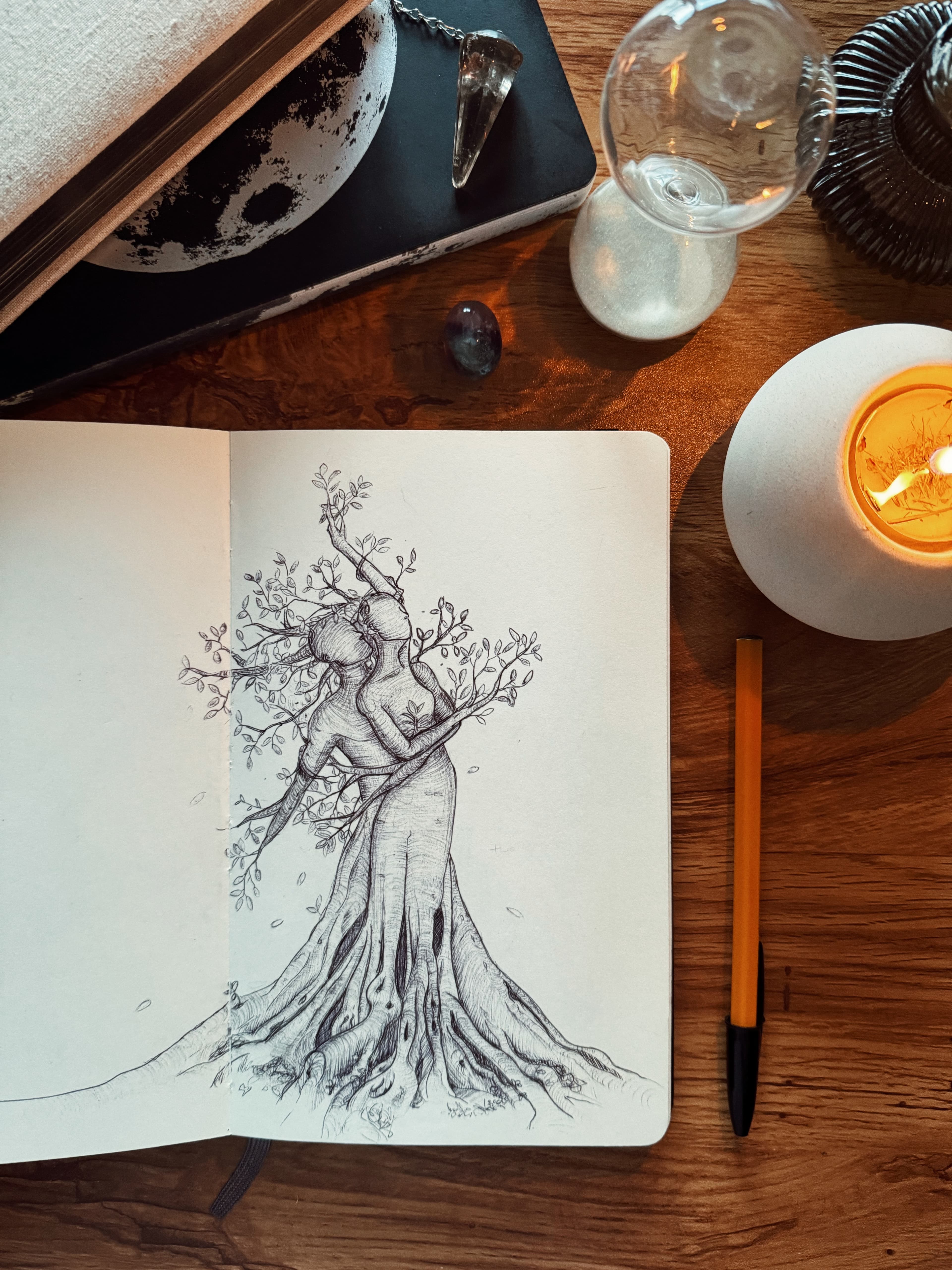 My sketchbook drawing of two intertwined figures with leafy branches and spreading roots, on a wooden table beside a pen and a lit candle.